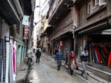 Narrow street lined with fabric shops and pedestrians walking by. Various fabrics are displayed on the left side of the street, while several people walk past closed shop fronts. Overhead, numerous electrical wires crisscross between buildings, casting shadows on the textured surfaces of the brick and concrete structures.