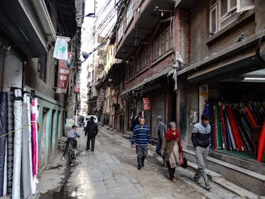 Narrow street lined with fabric shops and pedestrians walking by. Various fabrics are displayed on the left side of the street, while several people walk past closed shop fronts. Overhead, numerous electrical wires crisscross between buildings, casting shadows on the textured surfaces of the brick and concrete structures.