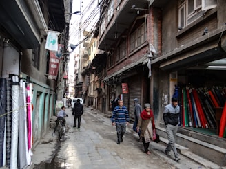 Narrow street lined with fabric shops and pedestrians walking by. Various fabrics are displayed on the left side of the street, while several people walk past closed shop fronts. Overhead, numerous electrical wires crisscross between buildings, casting shadows on the textured surfaces of the brick and concrete structures.