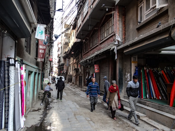 Narrow street lined with fabric shops and pedestrians walking by. Various fabrics are displayed on the left side of the street, while several people walk past closed shop fronts. Overhead, numerous electrical wires crisscross between buildings, casting shadows on the textured surfaces of the brick and concrete structures.