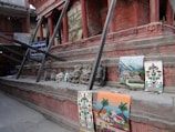 A series of colorful traditional masks and woven tapestries are displayed on the steps of a brick structure. The brick structure features intricate designs, and the tapestries depict various scenes including nature and flowers. Wooden beams support the structure, and a sign for a traditional mandala art school is visible in the background.