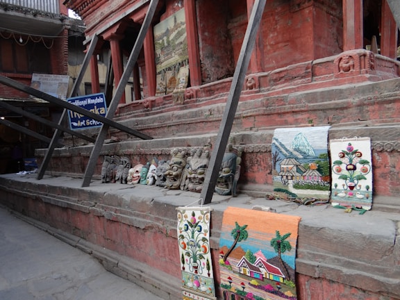 A series of colorful traditional masks and woven tapestries are displayed on the steps of a brick structure. The brick structure features intricate designs, and the tapestries depict various scenes including nature and flowers. Wooden beams support the structure, and a sign for a traditional mandala art school is visible in the background.