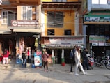 A busy urban street scene featuring a row of small shops, including a cafe and a spice and tea store. People walk on the sidewalk, some browsing the store displays. The shop signs are colorful and prominent, and various goods, like packaged snacks and hanging decorations, are visible. Motorbikes are parked along the street, and advertisements for treks and rentals adorn the building facades.
