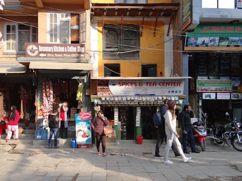 A busy urban street scene featuring a row of small shops, including a cafe and a spice and tea store. People walk on the sidewalk, some browsing the store displays. The shop signs are colorful and prominent, and various goods, like packaged snacks and hanging decorations, are visible. Motorbikes are parked along the street, and advertisements for treks and rentals adorn the building facades.