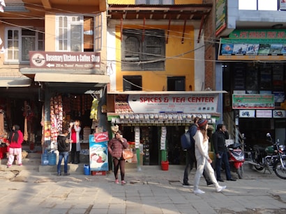 A busy urban street scene featuring a row of small shops, including a cafe and a spice and tea store. People walk on the sidewalk, some browsing the store displays. The shop signs are colorful and prominent, and various goods, like packaged snacks and hanging decorations, are visible. Motorbikes are parked along the street, and advertisements for treks and rentals adorn the building facades.