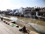 A riverside setting with a ceremonial atmosphere; smoke rises from several burning pyres along the riverbank. People are engaged in rituals, with offerings spread out on mats and steps leading down to the muddy river. Traditional architecture lines the opposite bank, with structures featuring domed roofs.