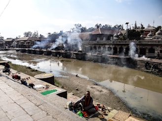 A riverside setting with a ceremonial atmosphere; smoke rises from several burning pyres along the riverbank. People are engaged in rituals, with offerings spread out on mats and steps leading down to the muddy river. Traditional architecture lines the opposite bank, with structures featuring domed roofs.