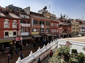A vibrant street scene in a bustling urban area features colorful buildings with various signs, including one for a tattoo shop. The architecture is traditional with red and orange facades, and numerous small shops line the street. People walk along the busy pathways, and greenery decorates the foreground.