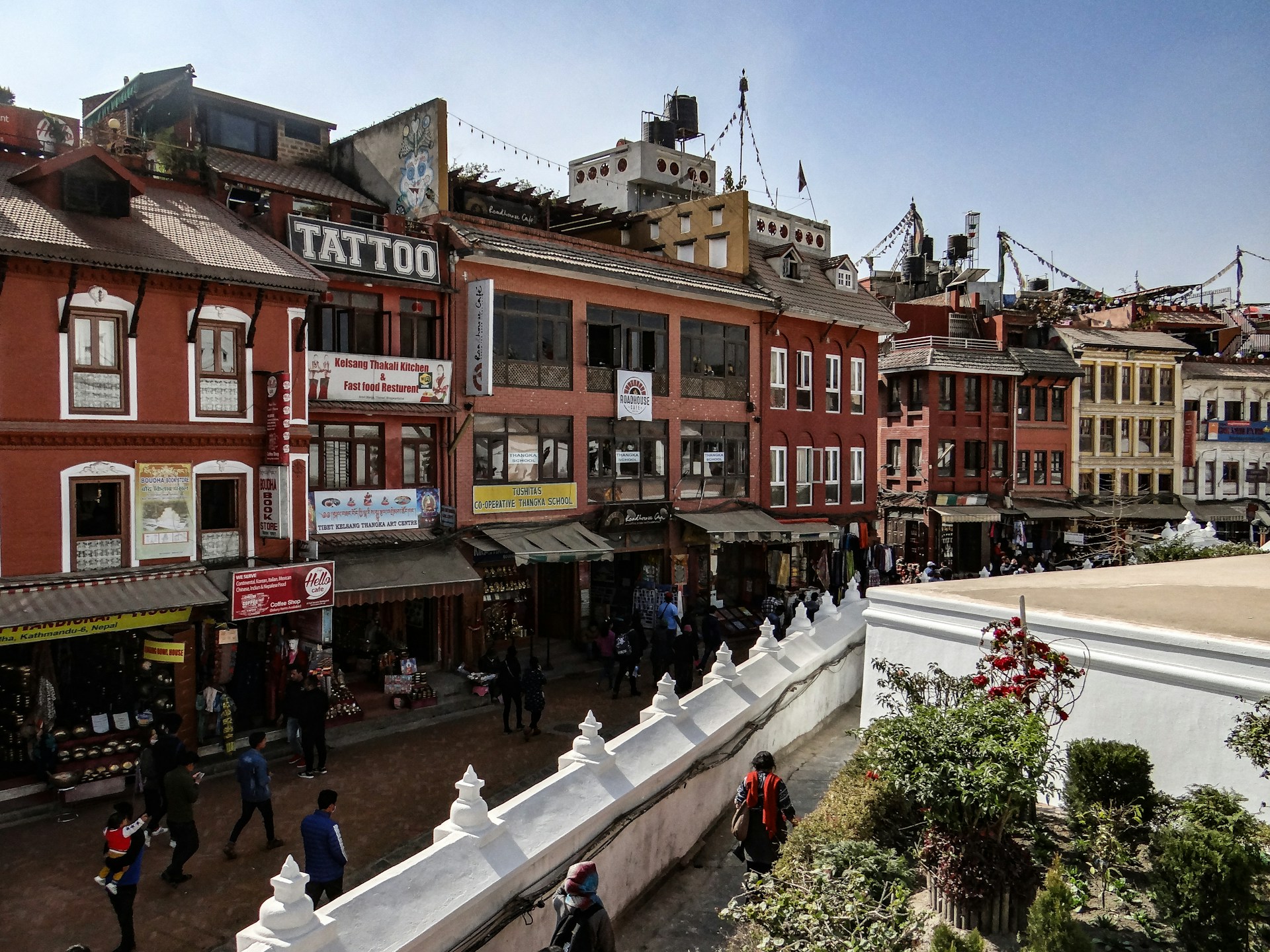 A vibrant street scene in a bustling urban area features colorful buildings with various signs, including one for a tattoo shop. The architecture is traditional with red and orange facades, and numerous small shops line the street. People walk along the busy pathways, and greenery decorates the foreground.