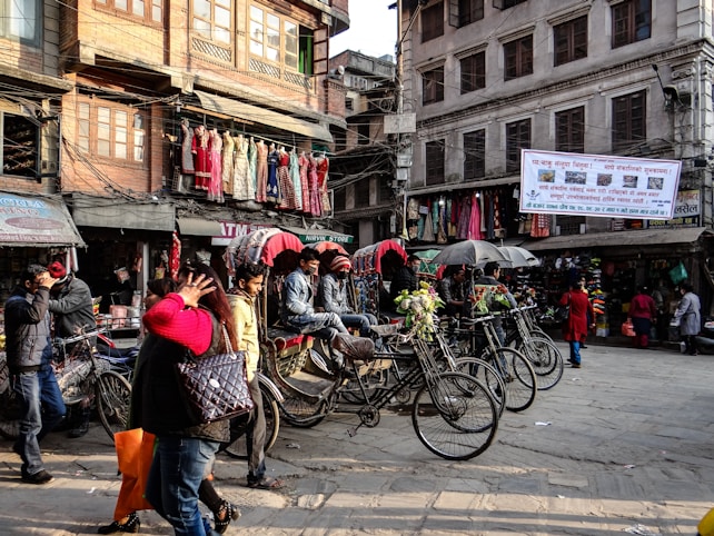 A busy street scene is captured with people walking and bicycles parked in the foreground. Vibrant fabrics and colorful garments hang on display outside a shop in the background. The architecture suggests an urban setting with multi-story buildings and banners displayed overhead. The environment appears lively and bustling.