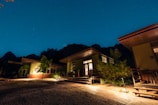 Several modern cabins with large windows and patios are illuminated at night. The scene is set under a clear, starry sky with mountains in the background. Trees and shrubs are planted around the buildings, and the ground is covered with gravel and concrete pathways.