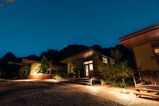 Several modern cabins with large windows and patios are illuminated at night. The scene is set under a clear, starry sky with mountains in the background. Trees and shrubs are planted around the buildings, and the ground is covered with gravel and concrete pathways.