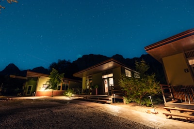 Several modern cabins with large windows and patios are illuminated at night. The scene is set under a clear, starry sky with mountains in the background. Trees and shrubs are planted around the buildings, and the ground is covered with gravel and concrete pathways.