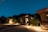 Several modern cabins with large windows and patios are illuminated at night. The scene is set under a clear, starry sky with mountains in the background. Trees and shrubs are planted around the buildings, and the ground is covered with gravel and concrete pathways.