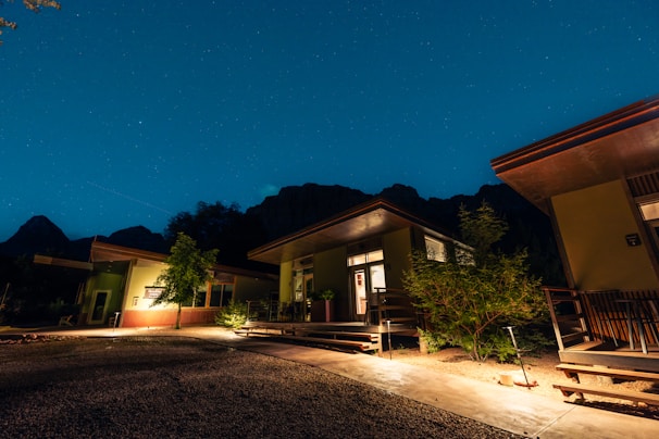 Several modern cabins with large windows and patios are illuminated at night. The scene is set under a clear, starry sky with mountains in the background. Trees and shrubs are planted around the buildings, and the ground is covered with gravel and concrete pathways.