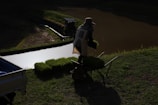 A person wearing a hat and protective clothing is carrying trays of seedlings next to a small body of water. The setting appears to be a rural, agricultural area with green grass surrounding the water's edge.