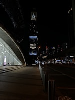 A nighttime urban scene with a modern architectural structure on the left featuring large glass windows and a softly lit interior. In the background, a tall tower with illuminated signage reading 'Sobha Realty' stands out against the dark sky. Street lights and distant skyscrapers enhance the cityscape, while the road is framed by stainless steel bollards.