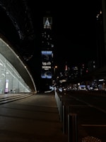 A nighttime urban scene with a modern architectural structure on the left featuring large glass windows and a softly lit interior. In the background, a tall tower with illuminated signage reading 'Sobha Realty' stands out against the dark sky. Street lights and distant skyscrapers enhance the cityscape, while the road is framed by stainless steel bollards.