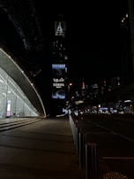 A nighttime urban scene with a modern architectural structure on the left featuring large glass windows and a softly lit interior. In the background, a tall tower with illuminated signage reading 'Sobha Realty' stands out against the dark sky. Street lights and distant skyscrapers enhance the cityscape, while the road is framed by stainless steel bollards.