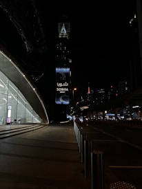 A nighttime urban scene with a modern architectural structure on the left featuring large glass windows and a softly lit interior. In the background, a tall tower with illuminated signage reading 'Sobha Realty' stands out against the dark sky. Street lights and distant skyscrapers enhance the cityscape, while the road is framed by stainless steel bollards.