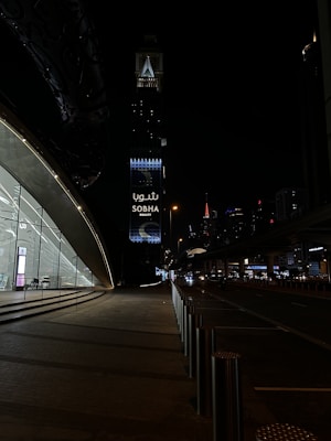 A nighttime urban scene with a modern architectural structure on the left featuring large glass windows and a softly lit interior. In the background, a tall tower with illuminated signage reading 'Sobha Realty' stands out against the dark sky. Street lights and distant skyscrapers enhance the cityscape, while the road is framed by stainless steel bollards.