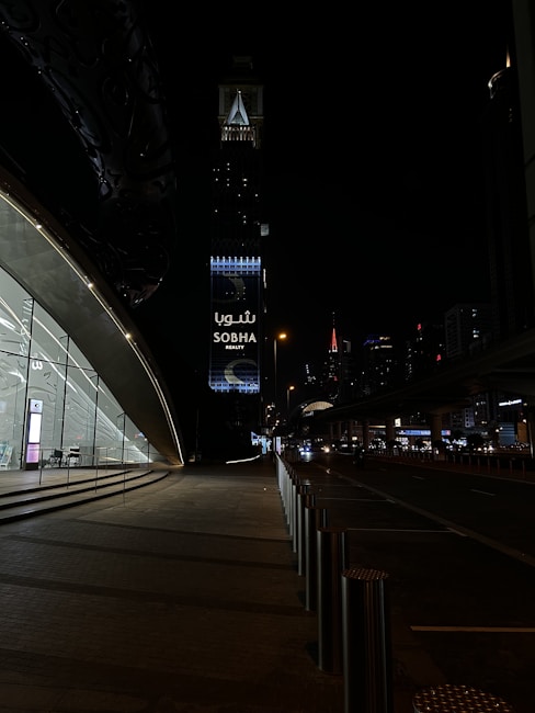 A nighttime urban scene with a modern architectural structure on the left featuring large glass windows and a softly lit interior. In the background, a tall tower with illuminated signage reading 