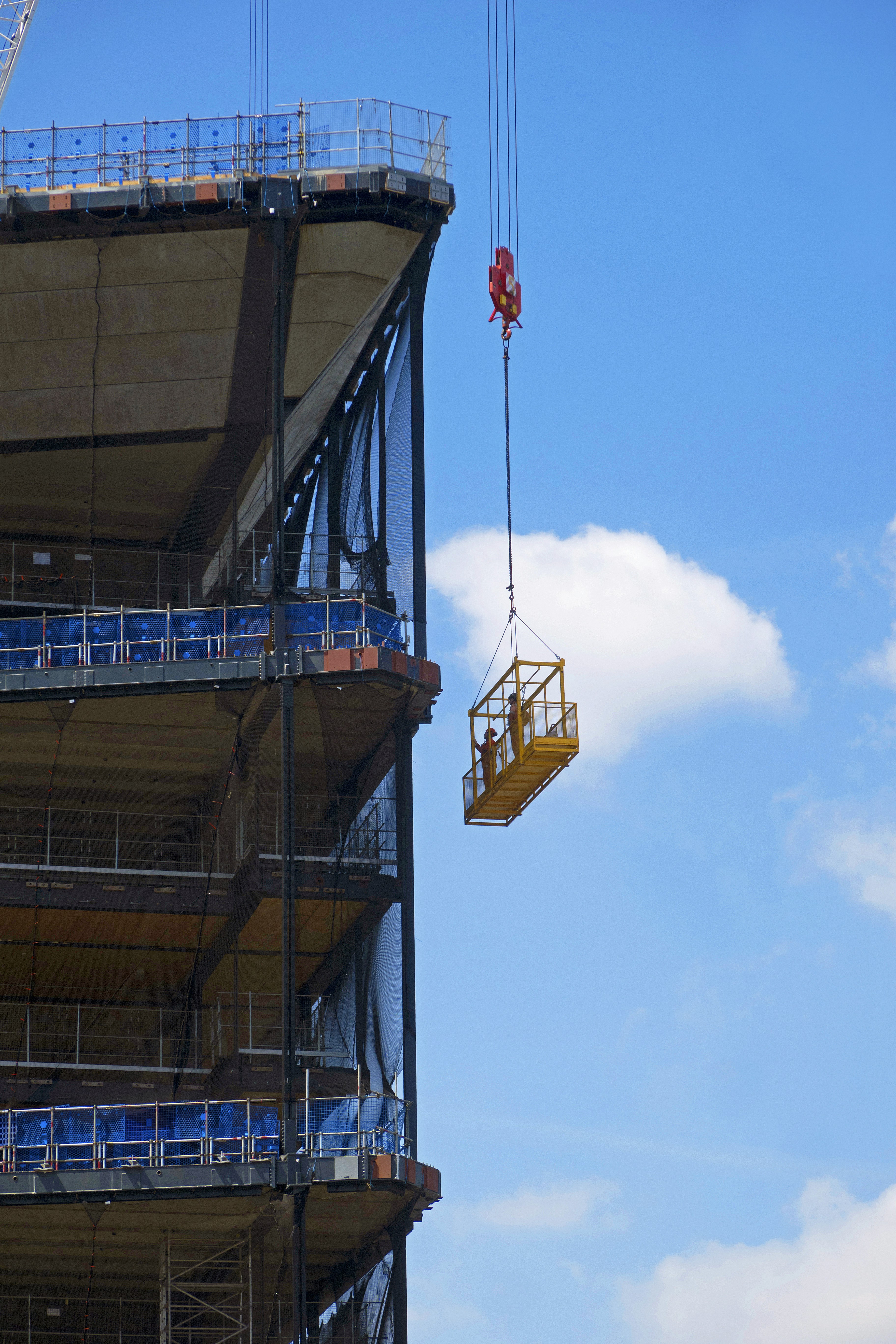 A construction elevator cage is suspended by a crane alongside a partially built multi-story structure. The clear blue sky and a few clouds are visible in the background. A worker is inside the cage, appearing to be at a significant height above the ground.