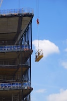 A construction elevator cage is suspended by a crane alongside a partially built multi-story structure. The clear blue sky and a few clouds are visible in the background. A worker is inside the cage, appearing to be at a significant height above the ground.