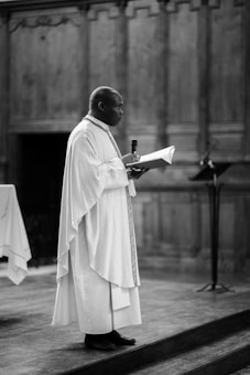 A person dressed in religious robes stands on a wooden platform, holding a microphone in one hand and a book in the other. The background consists of ornate wood paneling, suggesting a formal or religious setting.