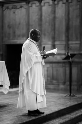 A person dressed in religious robes stands on a wooden platform, holding a microphone in one hand and a book in the other. The background consists of ornate wood paneling, suggesting a formal or religious setting.
