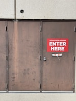 A set of three large brown metal doors with a noticeable sign on the middle door that reads 'Outdoor Centre Customers Enter Here.' The doors show signs of wear and use, with visible scratches and marks. Above the doors is a textured concrete wall.
