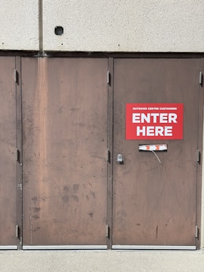 A set of three large brown metal doors with a noticeable sign on the middle door that reads 'Outdoor Centre Customers Enter Here.' The doors show signs of wear and use, with visible scratches and marks. Above the doors is a textured concrete wall.