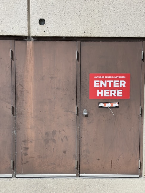 A set of three large brown metal doors with a noticeable sign on the middle door that reads 'Outdoor Centre Customers Enter Here.' The doors show signs of wear and use, with visible scratches and marks. Above the doors is a textured concrete wall.