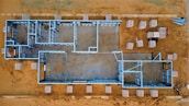 Aerial view of a construction site featuring an outlined architectural framework on a sandy ground. Metal beams form the structure of a building, with various rooms and hallways visible in the layout. Surrounding the framework are stacks of building materials organized on the ground.
