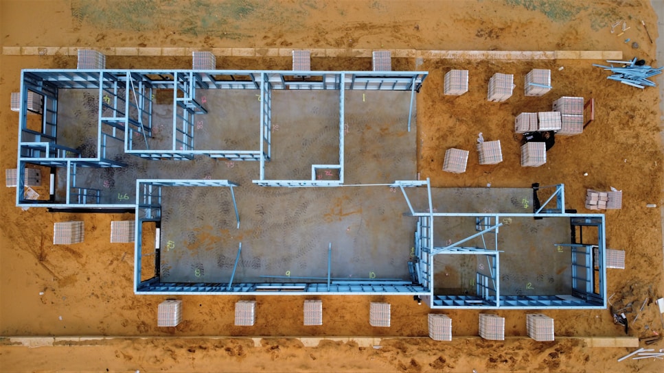 Aerial view of a construction site featuring an outlined architectural framework on a sandy ground. Metal beams form the structure of a building, with various rooms and hallways visible in the layout. Surrounding the framework are stacks of building materials organized on the ground.