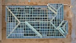 An aerial view of a construction site with a large rectangular structure framework. The grid-like pattern consists of intersecting beams and open spaces, giving it an intricate geometric appearance. The surrounding area is mostly dirt with some construction materials visible.