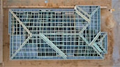 An aerial view of a construction site with a large rectangular structure framework. The grid-like pattern consists of intersecting beams and open spaces, giving it an intricate geometric appearance. The surrounding area is mostly dirt with some construction materials visible.