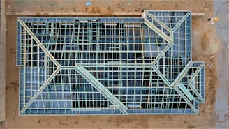 An aerial view of a construction site with a large rectangular structure framework. The grid-like pattern consists of intersecting beams and open spaces, giving it an intricate geometric appearance. The surrounding area is mostly dirt with some construction materials visible.