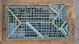 An aerial view of a construction site with a large rectangular structure framework. The grid-like pattern consists of intersecting beams and open spaces, giving it an intricate geometric appearance. The surrounding area is mostly dirt with some construction materials visible.