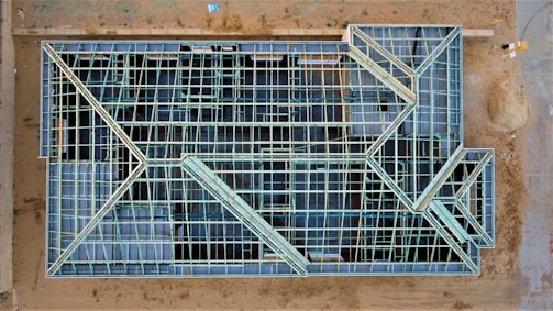 An aerial view of a construction site with a large rectangular structure framework. The grid-like pattern consists of intersecting beams and open spaces, giving it an intricate geometric appearance. The surrounding area is mostly dirt with some construction materials visible.