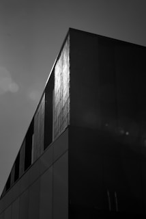 A monochrome architectural photograph of a modern building with a geometric design. The structure features clean lines and a smooth facade with sunlight reflecting off its surface, creating a stark contrast of light and shadow.
