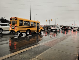 A small yellow school bus is traveling on a wet road during a rainy day. The surrounding environment includes multiple vehicles waiting in traffic, with their headlights and brake lights reflecting on the wet surface. The sky is overcast, and the overall scene conveys a dreary, rainy day.