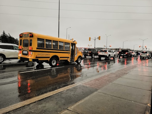 A small yellow school bus is traveling on a wet road during a rainy day. The surrounding environment includes multiple vehicles waiting in traffic, with their headlights and brake lights reflecting on the wet surface. The sky is overcast, and the overall scene conveys a dreary, rainy day.