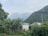 A lush, green landscape featuring tall banana plants and dense foliage in the foreground. A small tin-roofed structure is nestled among the greenery. In the background, majestic mountains rise under a cloudy sky, creating a serene and natural setting.
