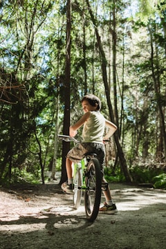 A young person rides a bicycle along a sunlit path in a dense forest, surrounded by tall trees and lush greenery. Sunlight filters through the canopy, casting dappled shadows on the ground.