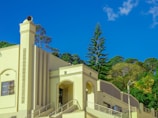 A building with Art Deco architectural elements, featuring sleek geometric lines and a prominent column with a speaker on top. The structure is painted in a light yellow hue, contrasting with the clear blue sky and lush green trees in the background. There are metal railings and a metal cylinder visible beside the building.