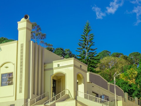 A building with Art Deco architectural elements, featuring sleek geometric lines and a prominent column with a speaker on top. The structure is painted in a light yellow hue, contrasting with the clear blue sky and lush green trees in the background. There are metal railings and a metal cylinder visible beside the building.