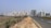 A long, straight road flanked by a dirt path and tall, modern apartment buildings in the background. Electrical poles with cables run along the side of the road. The area on one side of the road is grassy with shrubs while the other side has construction and multiple high-rise buildings.