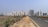 A long, straight road flanked by a dirt path and tall, modern apartment buildings in the background. Electrical poles with cables run along the side of the road. The area on one side of the road is grassy with shrubs while the other side has construction and multiple high-rise buildings.