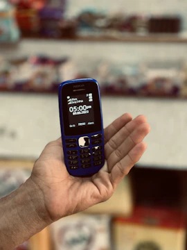 A hand holding a blue Nokia feature phone, displaying the time and date on the screen. The background appears to be a blurred setting with various items on shelves.